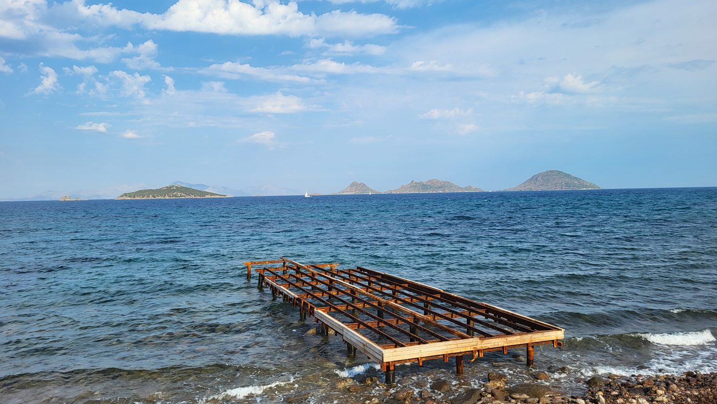 A half-built wooden pier reaching into a blue sea, low islands on the horizon under scattered cloud.