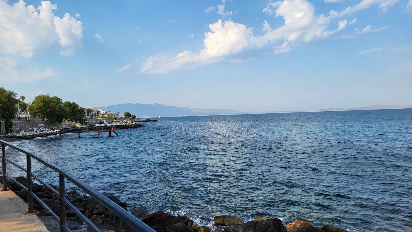 A seafront railing above dark rocks, a calm bay and distant mountains under a clouded sky.